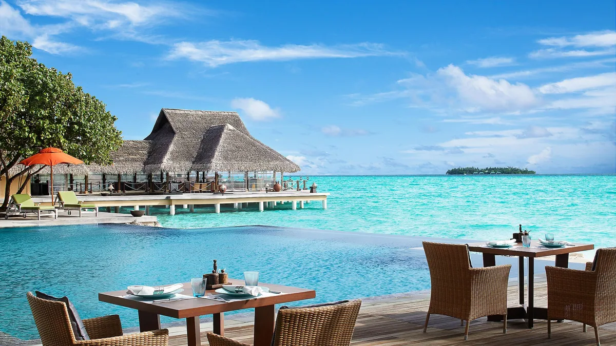 Taj Exotica Resort & Spa, Maldives, dining, South Malé Atoll, Maldives, Principal pavilion exterior at midday, twin thatched peaks rising above the over-water dining deck, an infinity-edge pool in the foreground, wicker dining tables on the timber boardwalk with a distant island on the horizon.