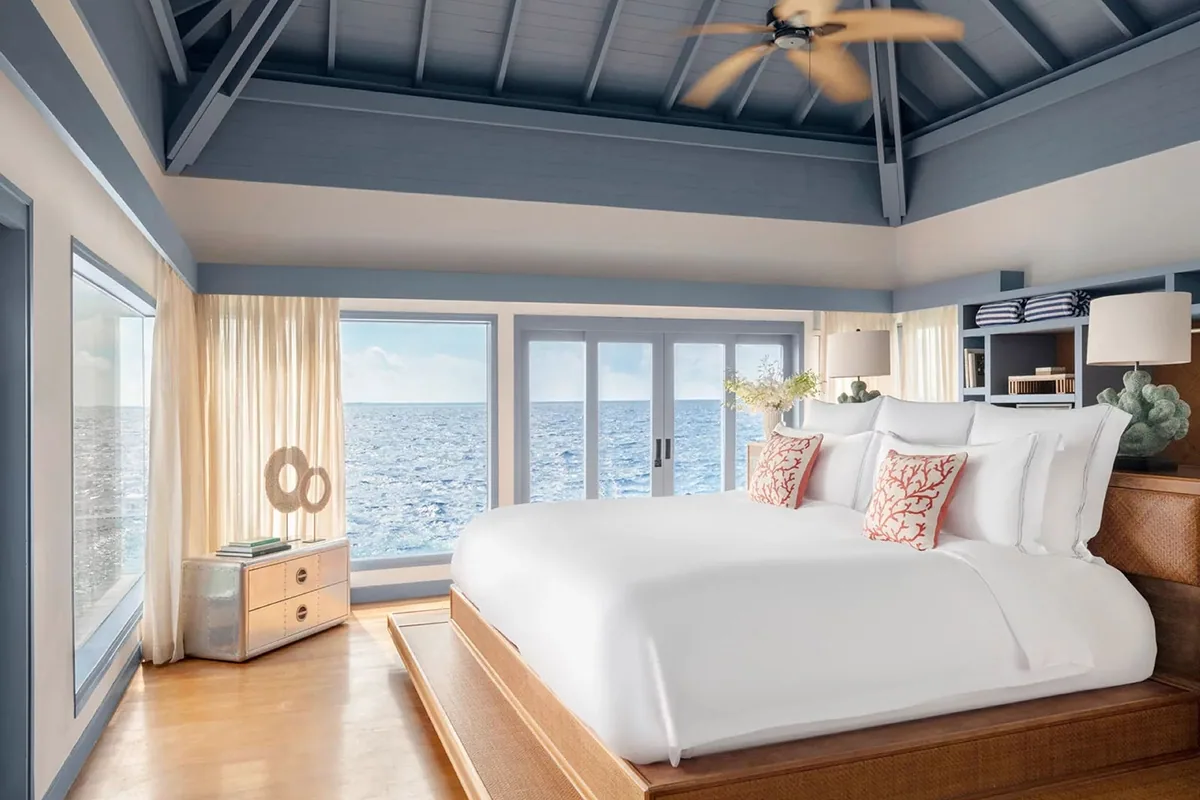 The Halcyon Private Isles Maldives, villa interior, Gaafu Alifu Atoll, Maldives, Overwater Villa bedroom, the property's signature vaulted pale-blue beamed ceiling with ceiling fan, king bed with white linens and coral-pattern accent pillows, twin glass sliders opening to the deep-ocean horizon, woven rattan headboard.