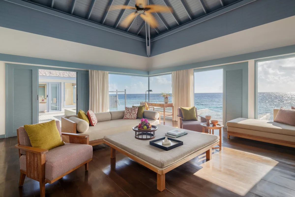 The Halcyon Private Isles Maldives, villa interior, Gaafu Alifu Atoll, Maldives, Overwater Villa living room, vaulted pale-blue ceiling and pale-blue louvered doors, beige modular sofa and daybed grouped around a low table, twin armchairs with yellow and pink accent cushions (Halcyon-era updated palette), glass sliders to the deck and ocean.