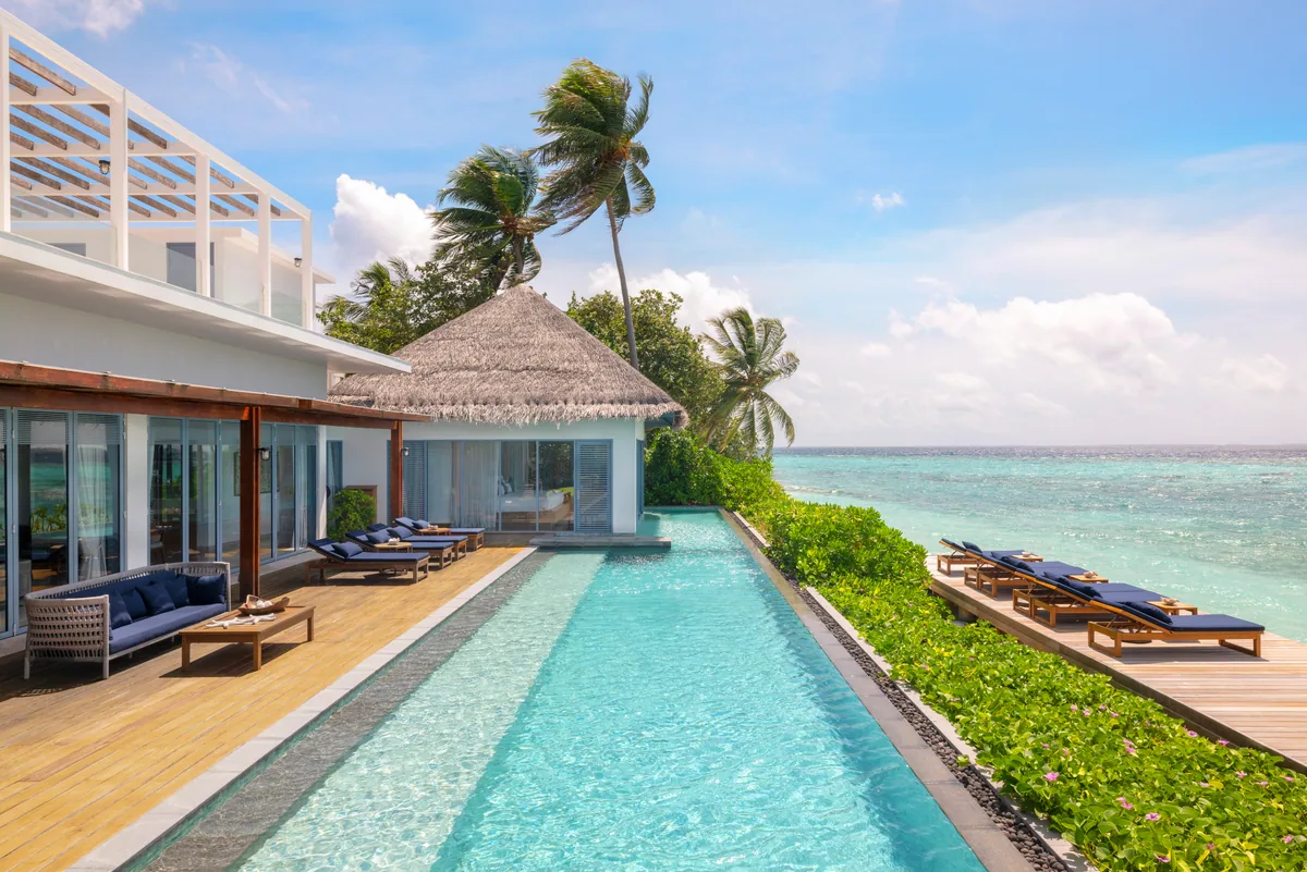 The Halcyon Private Isles Maldives, villa beach, Gaafu Alifu Atoll, Maldives, Grand Estate private lap pool, a long narrow lap pool running flush along the reef edge with navy-cushion sun loungers on the timber deck on both sides, the white modernist villa with two-storey rooftop sundeck pavilion at left, palm canopy beyond.