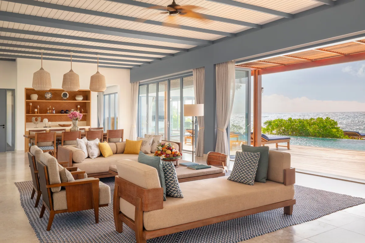 The Halcyon Private Isles Maldives, villa interior, Gaafu Alifu Atoll, Maldives, Grand Estate living room, large open-plan space with grey-blue beamed ceiling, twin daybed sofas and an L-shaped sofa with yellow and green accent cushions, woven rattan pendant trio over the dining table at left, glass sliders to the deck and lap pool and ocean.