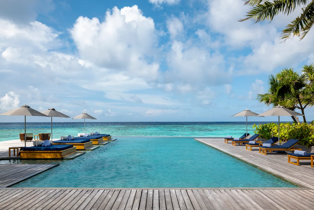 The Halcyon Private Isles Maldives, setting, Gaafu Alifu Atoll, Maldives, Main island infinity pool with twin floating sun-lounger pavilions flanking the centre of the pool with beige parasols, navy-striped sun loungers on the timber deck at the right, palm canopy framing, the lagoon stretching to the horizon at the infinity edge.