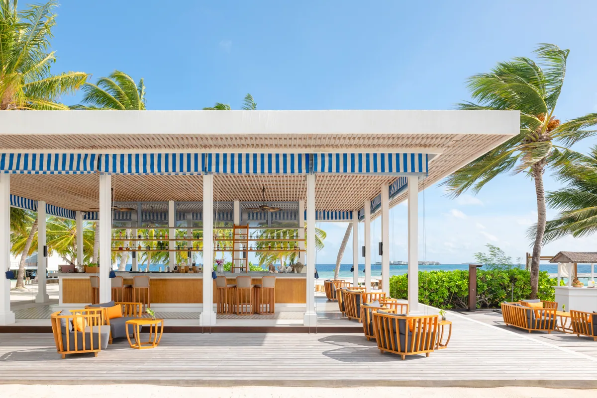 The Halcyon Private Isles Maldives, dining, Gaafu Alifu Atoll, Maldives, Bell Bar, the signature blue-and-white striped pergola roof over the open-air bar with white columns and bar counter with stools, orange-rope-back swivel chairs around timber tables on the deck, palm trees framing the beachside setting.