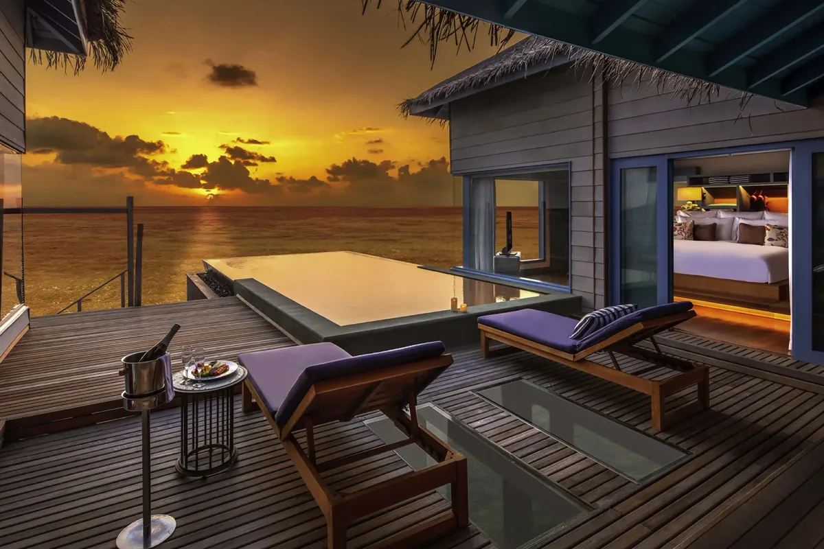 The Halcyon Private Isles Maldives, villa overwater, Gaafu Alifu Atoll, Maldives, Overwater Pool Suite at the over-water configuration with private pool: sunset-orientation timber deck, infinity-edge plunge pool, plunge into the lagoon at the deck edge, palm-thatch eaves, and the open-plan bedroom visible through the floor-to-ceiling glazing.