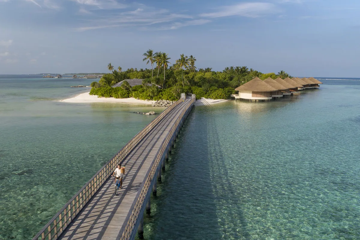 The Residence Maldives at Dhigurah, setting, Gaafu Alifu Atoll, Maldives, Signature long jetty-boardwalk bridge extending from the lagoon toward the palm-canopy island, a couple cycling toward the island, the over-water villa row visible at right, deep-turquoise lagoon stretching with the sister island visible in the distance.