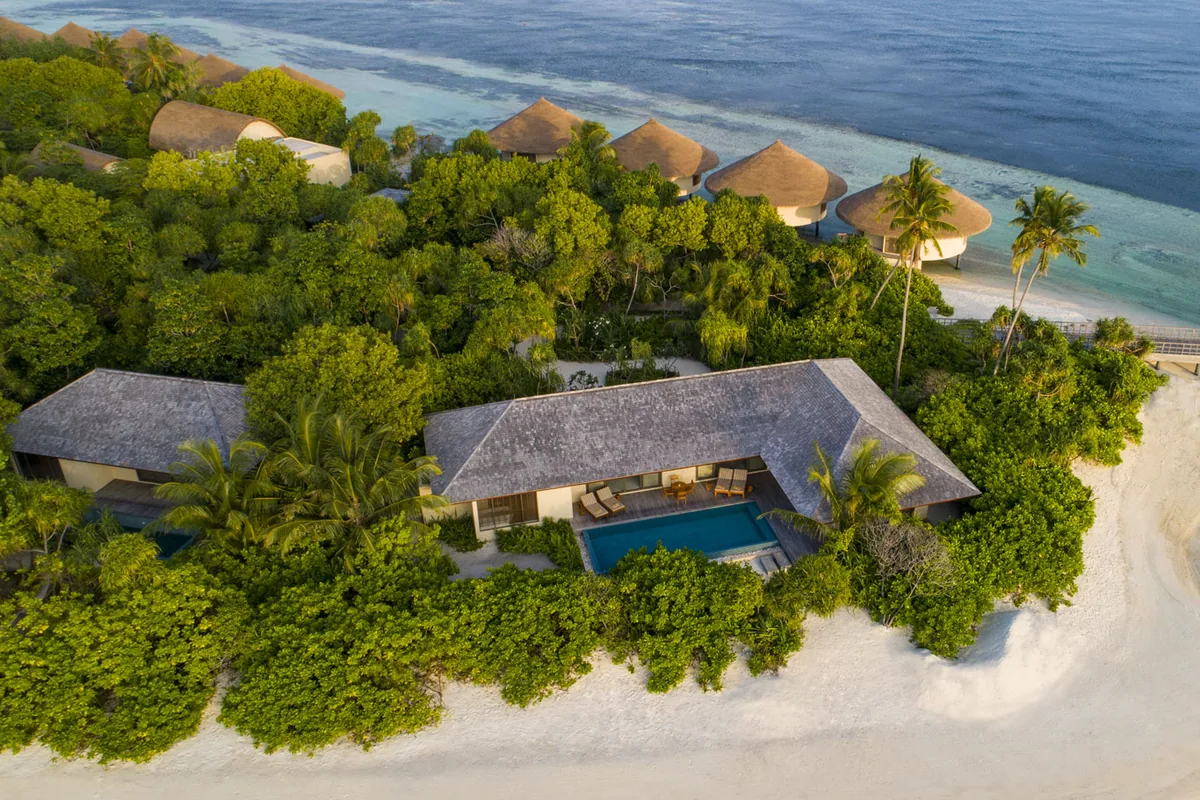 The Residence Maldives at Dhigurah, aerial, Gaafu Alifu Atoll, Maldives, Aerial of the Beach Pool Villa cluster, palm-canopy island with the white-sand beach perimeter, smaller round-roof villas above the larger gabled Beach Pool Villas, reef gradient from turquoise shallows to deep blue at the cluster edge.