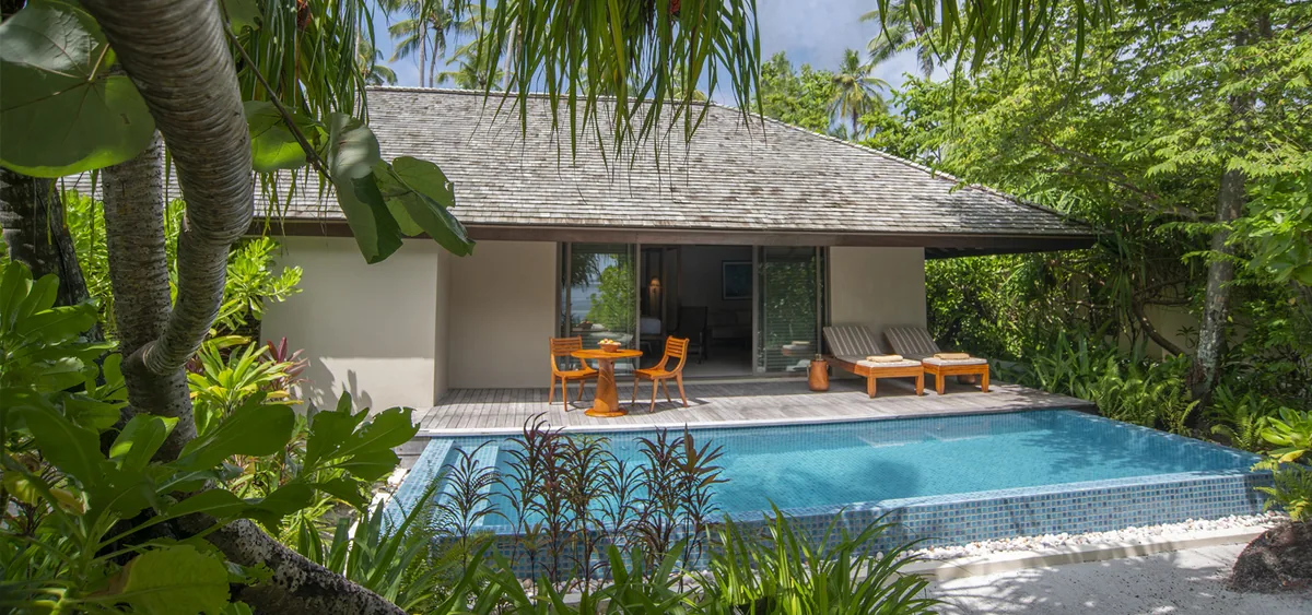 The Residence Maldives at Dhigurah, villa beach, Gaafu Alifu Atoll, Maldives, Beach Pool Villa exterior, modern grey-tile-roof villa with a private rectangular plunge pool and dining set on the timber deck, twin orange sun loungers, dense palm canopy framing the villa entry.