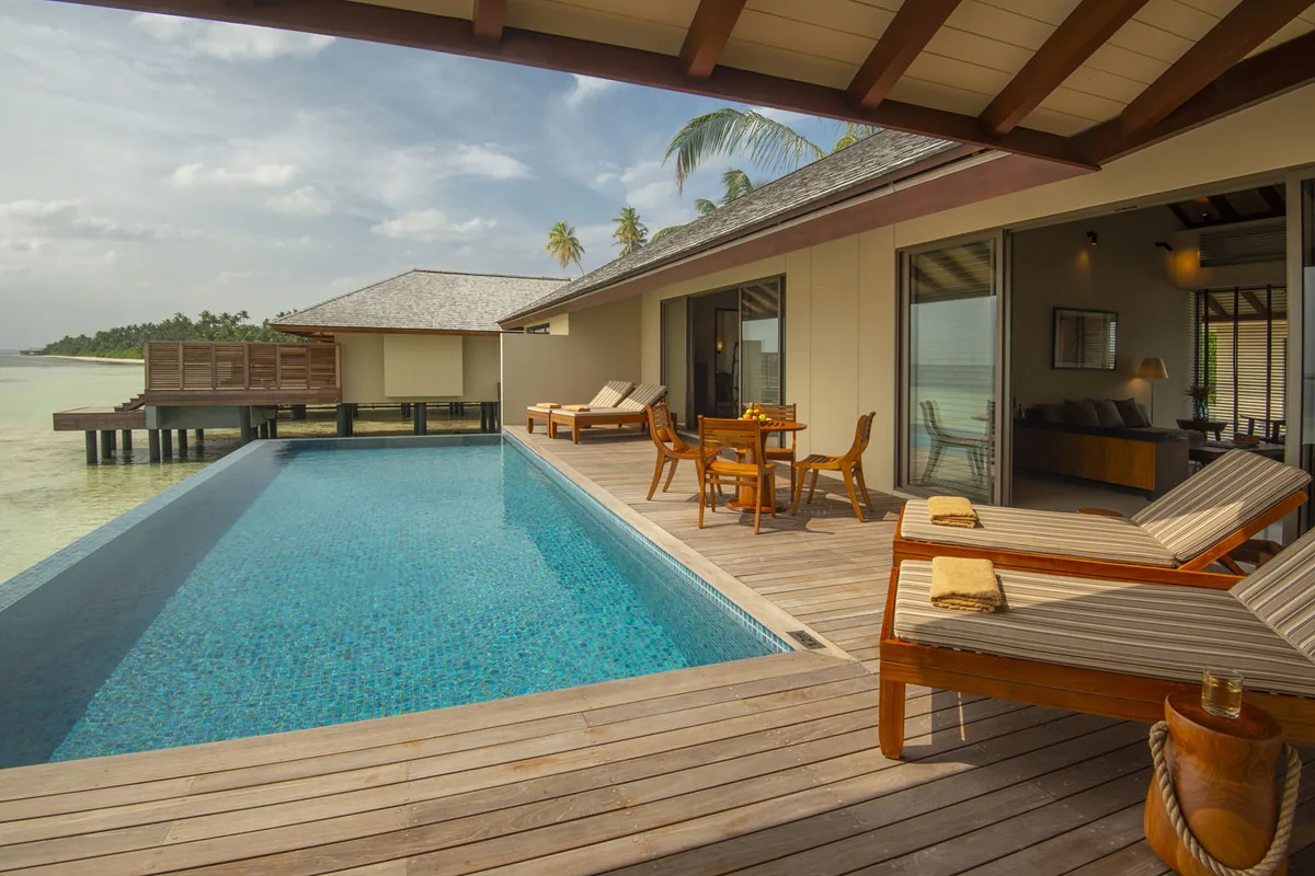 The Residence Maldives at Dhigurah, villa overwater, Gaafu Alifu Atoll, Maldives, Water Villa with Pool deck, a large mosaic-tile plunge pool with striped wood sun loungers on the timber deck, dining set, sliding doors opening to the villa interior at right, ocean view stretching beyond with a glimpse of the over-water Spa pavilion at left.