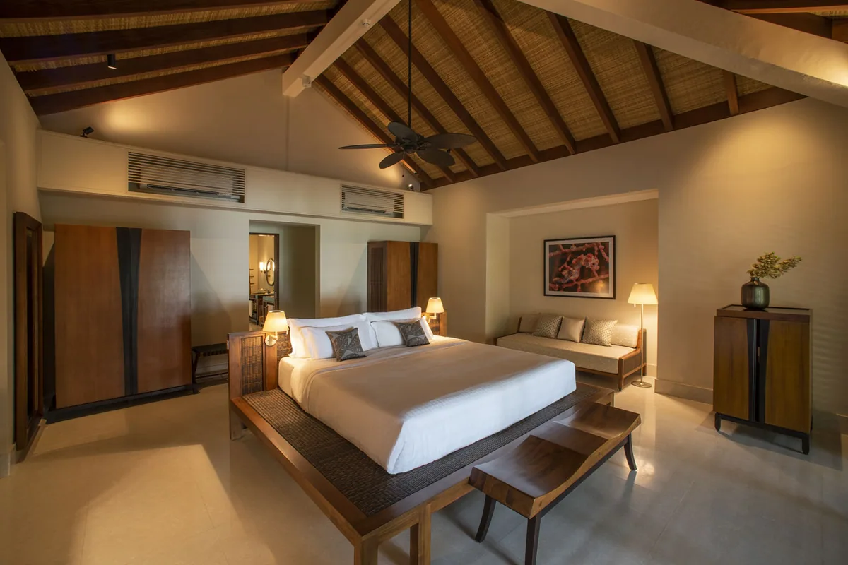 The Residence Maldives at Dhigurah, villa interior, Gaafu Alifu Atoll, Maldives, Villa bedroom interior, vaulted thatch-and-timber-beam ceiling with ceiling fan, dark-hardwood platform bed with white linens and grey accent pillows, woven-wood bench at the foot of the bed, daybed sofa with cushions, twin armoires and a dresser, framed coral-life artwork on the wall.