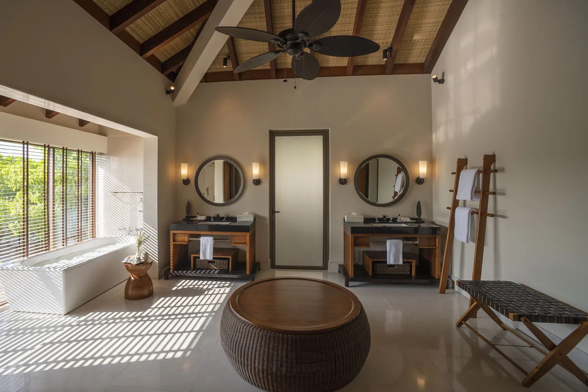 The Residence Maldives at Dhigurah, villa interior, Gaafu Alifu Atoll, Maldives, Villa bathroom with vaulted timber-and-thatch ceiling and ceiling fan, twin round-mirror vanities on dark-marble counters, a freestanding tub at left framed by bamboo blinds and a small woven side table, a woven-rattan ottoman in the centre, a classic-style timber towel ladder at right.
