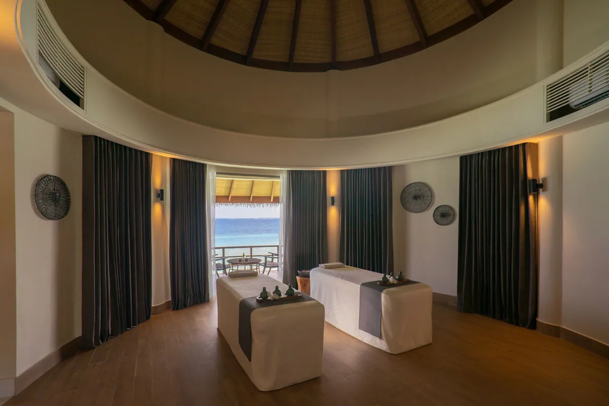 The Residence Maldives at Dhigurah, spa, Gaafu Alifu Atoll, Maldives, The Spa by Clarins overwater pavilion interior, circular dome ceiling with thatched cone, twin massage tables in the centre, dark velvet curtains pulled aside to reveal sliding doors opening onto an ocean-view deck with chairs and a daybed.