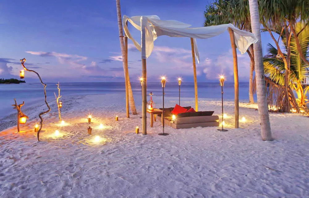 The Residence Maldives at Falhumaafushi, dining, Gaafu Alifu Atoll, Maldives, Romantic private beach dinner setup at twilight, a bamboo-pole canopy with draped sheer fabric over a candle-lit lounger nook with red accent pillows, bamboo torches and lantern-lit driftwood arrangements on the white-sand beach, the calm sea at the twilight horizon.