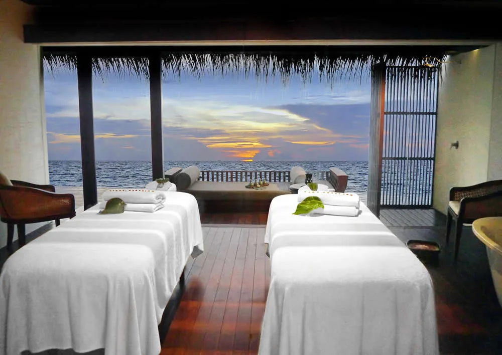 The Residence Maldives at Falhumaafushi, spa, Gaafu Alifu Atoll, Maldives, The Spa by Clarins overwater couple treatment room at sunset, twin massage tables with white linens and leaf accents on the dark hardwood floor, the open thatched-eave perimeter revealing the daybed terrace and golden-sky horizon over the lagoon.