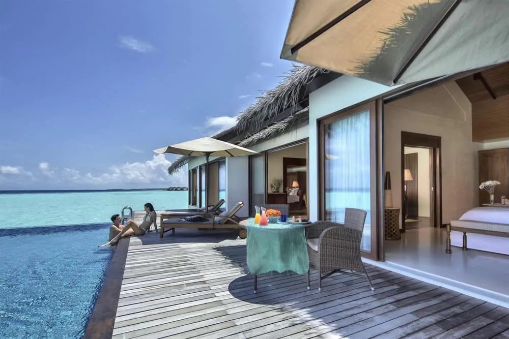 The Residence Maldives at Falhumaafushi, villa overwater, Gaafu Alifu Atoll, Maldives, Water Pool Villa deck, a couple at the infinity-edge pool with cocktails, the thatched-eave villa with twin striped-cushion sun loungers, dining set on the deck, open sliders to the bedroom interior with king bed visible, palm canopy at the horizon.