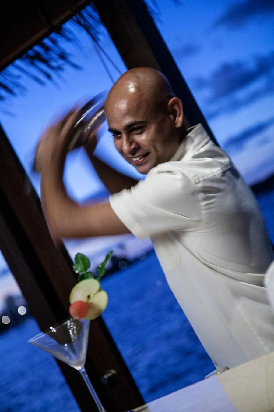 The Residence Maldives at Falhumaafushi, dining, Gaafu Alifu Atoll, Maldives, Bartender shaking cocktail at twilight at the beach bar, a garnished martini glass in the foreground, the open thatched-eave bar architecture and a blue-pink twilight lagoon backdrop with bokeh-lit perimeter.