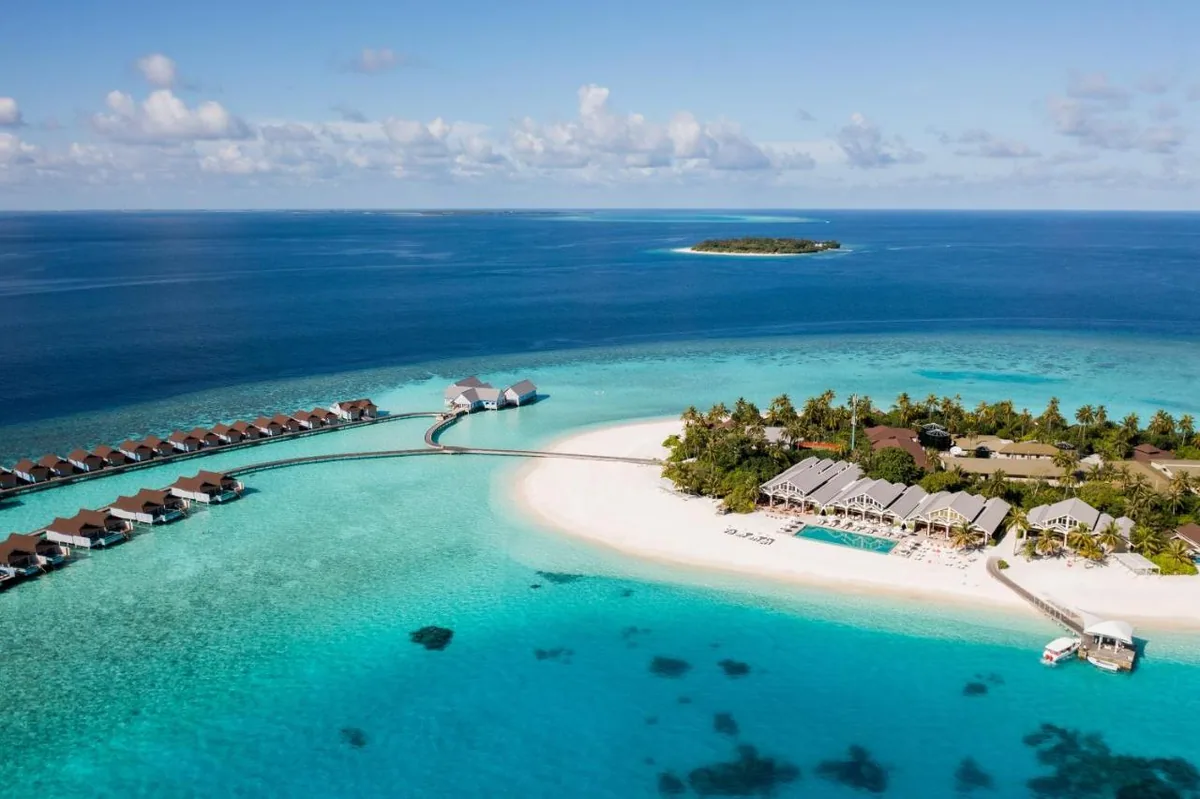 The Standard Maldives, aerial, Raa Atoll, Maldives, The Standard Huruvalhi drone aerial: two-row overwater villa configuration on stilts at left tracking along a curved boardwalk to a circular overwater pavilion (destination dining venue), main beach with crescent shape and beach-villa cluster + pool at centre-right, additional thatched buildings extending into the interior, small uninhabited islet in distance, deep teal-to-blue lagoon transitioning to ultramarine open ocean. The Huruvalhi island in north-central Raa Atoll with central-pool-deck daily-rhythm anchor.