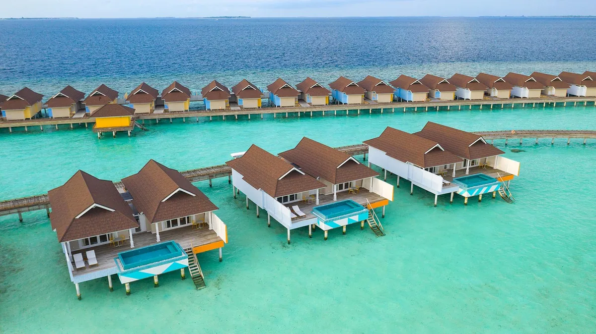 The Standard Maldives, villa overwater, Raa Atoll, Maldives, Overwater Villa with Pool cluster top-down aerial: triple grey-shingle pyramid-roof villas with white walls and ORANGE color-block accent panels, integrated cantilevered plunge pools with turquoise + blue + white GEOMETRIC TILE PATTERNS on each villa, and an extending OWV row in background with multi-color accents (orange, yellow, blue). The Standard brand vocabulary translated to the Maldivian lagoon-side layout.