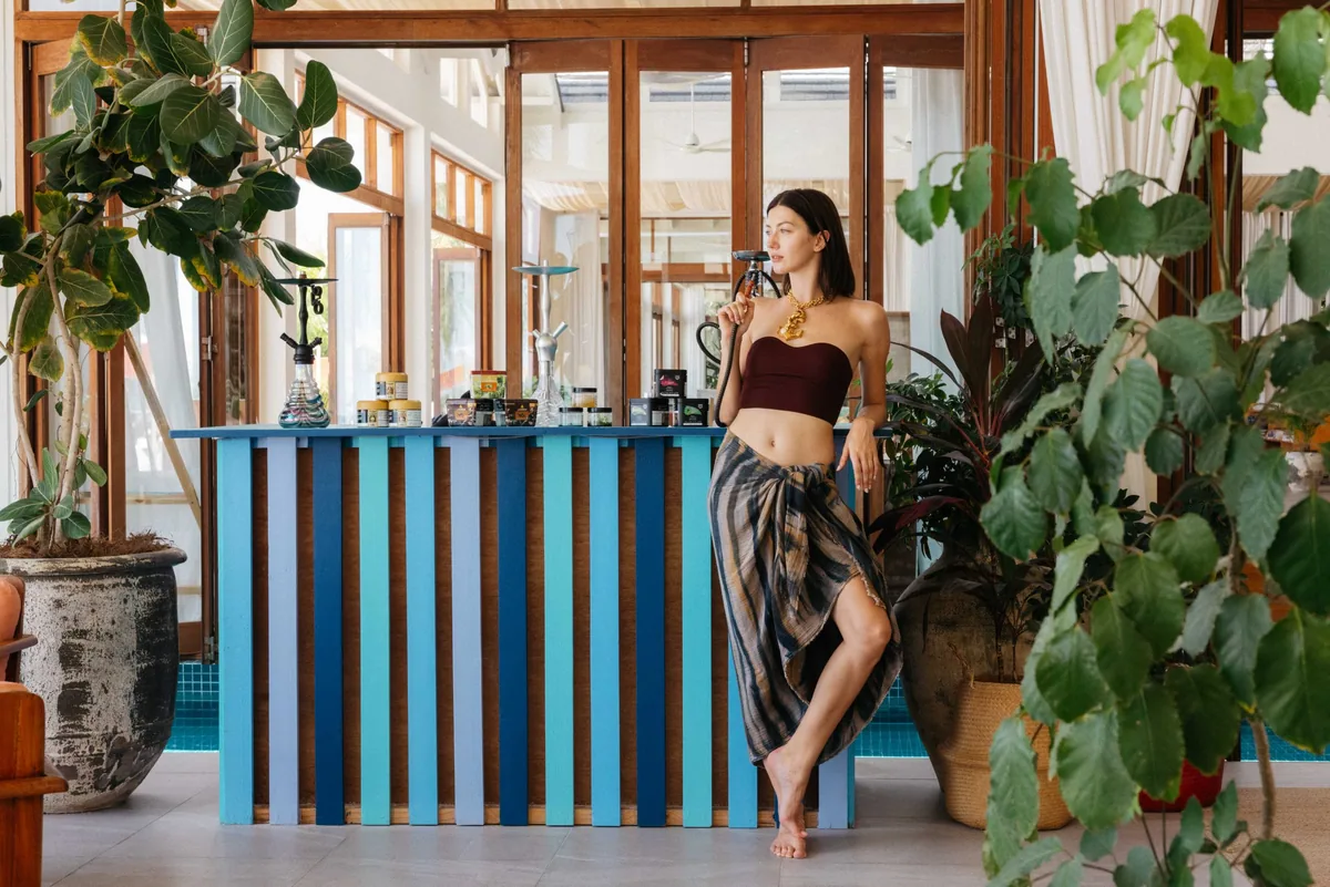 The Standard Maldives, dining, Raa Atoll, Maldives, Todis bar interior, open-air pavilion with timber-louvred panels and wide opening into the pool deck behind, distinctive vertical-stripe bar facade in graduated blues, teals and turquoise (four-tone wood-slat palette), bartender behind the counter, large fiddle-leaf-fig and banana-leaf plants framing the foreground. The structural food-and-beverage signature of the daily pool-side rhythm.