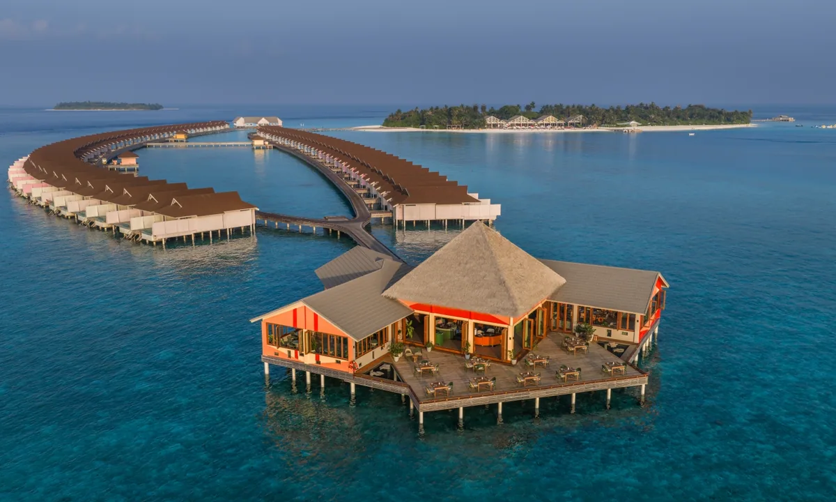 The Standard Maldives, dining, Raa Atoll, Maldives, Onda Mediterranean restaurant standalone overwater pavilion aerial, peaked thatched roof above the orange color-block facade (Standard brand DNA), open deck dining tables, anchored at the end of an access bridge reading back to the arced overwater villa row and the main island beach in the distance.