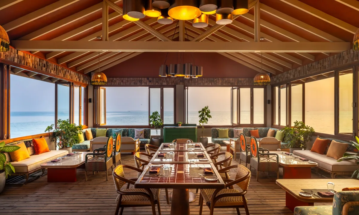The Standard Maldives, dining, Raa Atoll, Maldives, Onda interior, multi-disc bronze pendant chandelier suspended under the peaked timber-rafter ceiling, terracotta wood-slat backsplash, central long dining table surrounded by sunken-floor banquette seating in orange, green and cream cushion blocks, accent screens flanking, ocean horizon across the floor-to-ceiling window walls.