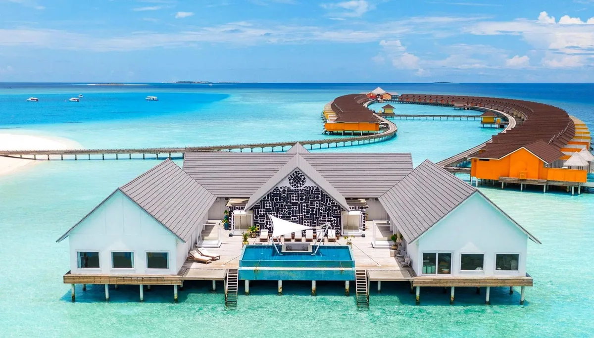 The Standard Maldives, spa, Raa Atoll, Maldives, Overwater Spa signature aerial, triple-pavilion structure with white-painted gable, black-pattern backsplash on the central pavilion, plunge pool, cushioned daybeds, arced orange-and-white overwater villa row curving across the background with grey-shingle pyramid roofs and clear turquoise lagoon. The on-property spa pavilion at the design-hotel wellness configuration.