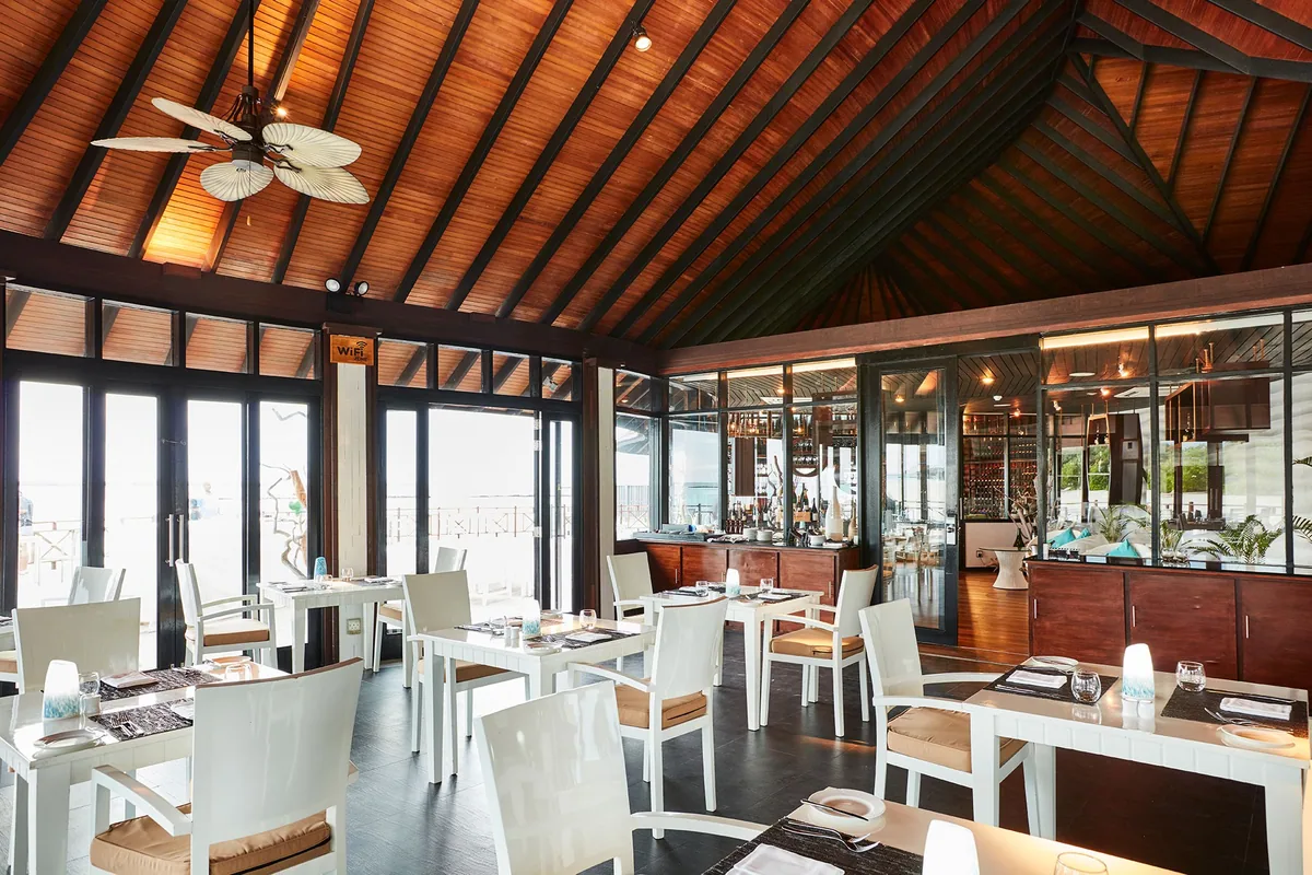 Sun Siyam Iru Fushi, Flavours French interior with cathedral wooden ceiling, palm-frond ceiling fan, white tables and chairs and glass walls onto lagoon, Noonu Atoll, Maldives