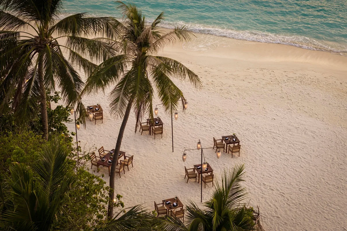 Sun Siyam Iru Fushi, Islander's Grill wooden tables and chairs on white sand under coconut palms with lantern posts and ocean lapping, Noonu Atoll, Maldives
