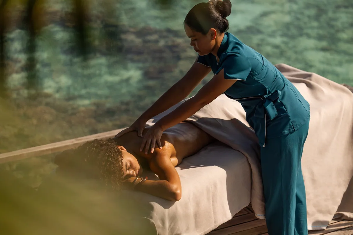 Sun Siyam Iru Fushi, tropical spa therapist in teal uniform applying back massage on overwater treatment table with shoreline coral visible through water, Noonu Atoll, Maldives