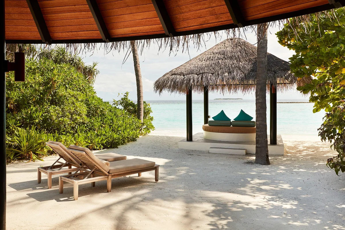 Sun Siyam Iru Fushi, Beach Villa porch view onto thatched daybed pavilion with teal cushions, sun loungers on white sand and lagoon, Noonu Atoll, Maldives