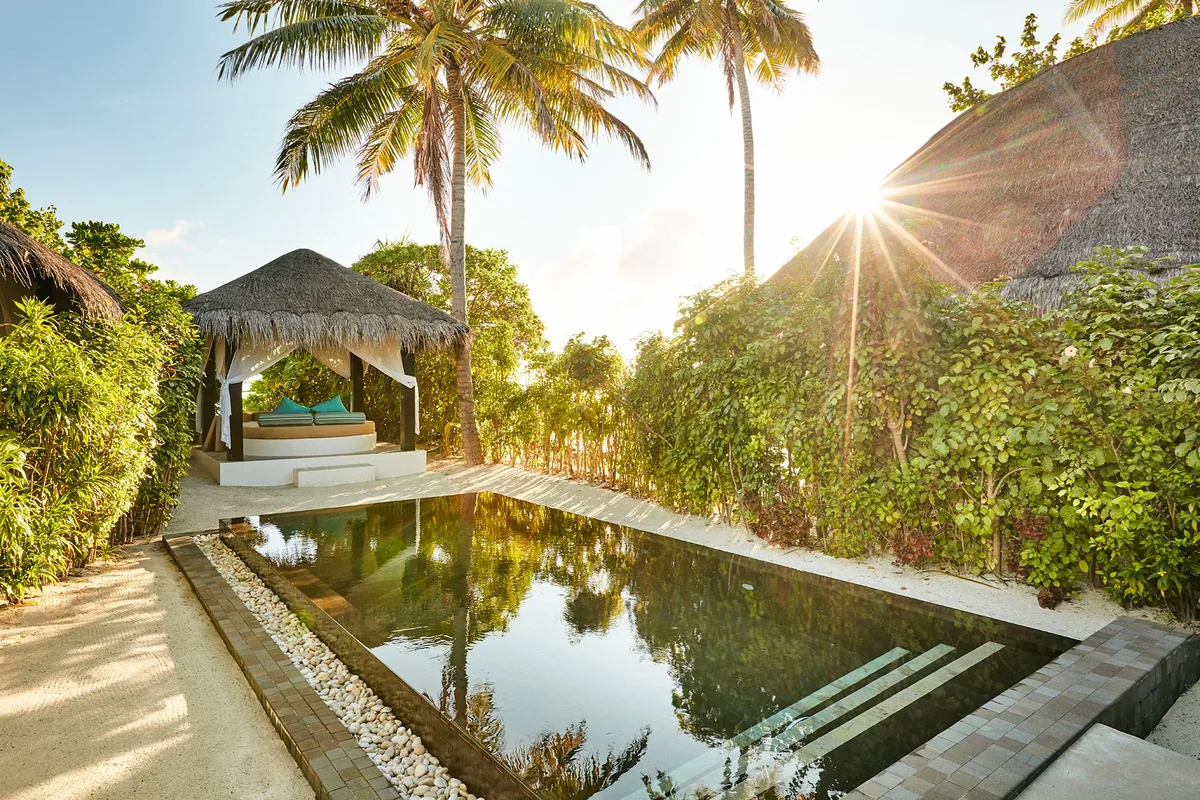 Sun Siyam Iru Fushi, Deluxe Beach Villa with Pool, thatched daybed pavilion next to dark-tile rectangular infinity pool with sunburst through palms, Noonu Atoll, Maldives