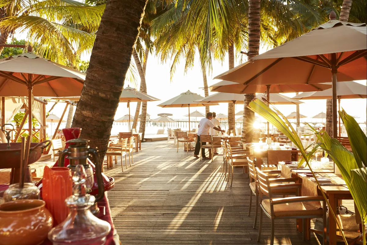 Sun Siyam Iru Fushi, Taste of India beachfront deck with parasol-covered wooden tables under coconut palms, decorative brassware and sunset light, Noonu Atoll, Maldives