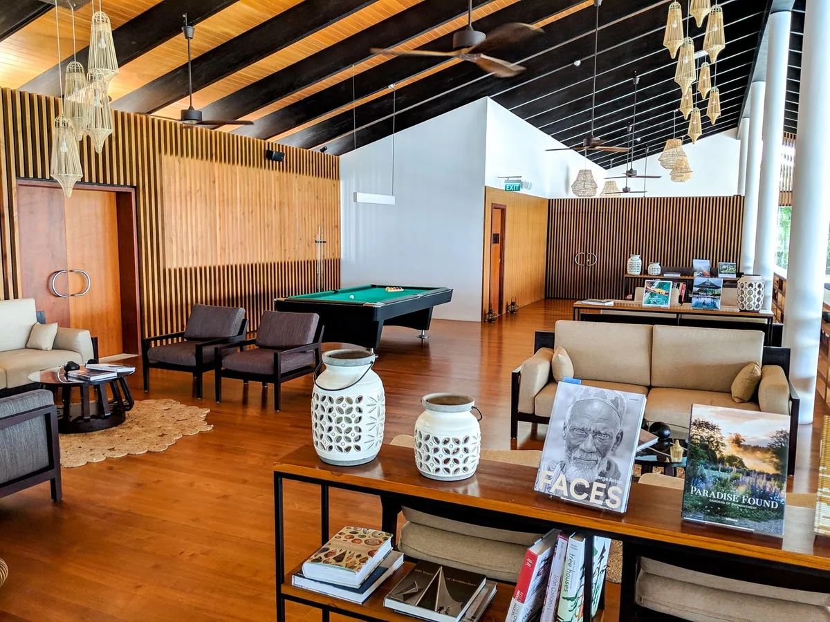 The Westin Maldives Miriandhoo Resort, setting, Baa Atoll, Maldives, Resort lounge and library, the warm-timber-floored room with the green-felt pool table at the back, mid-century-style lounge sofas, white ginger-jar table lamps, dark slatted-wood feature wall and the coffee-table books, including FACES, on the open shelves.