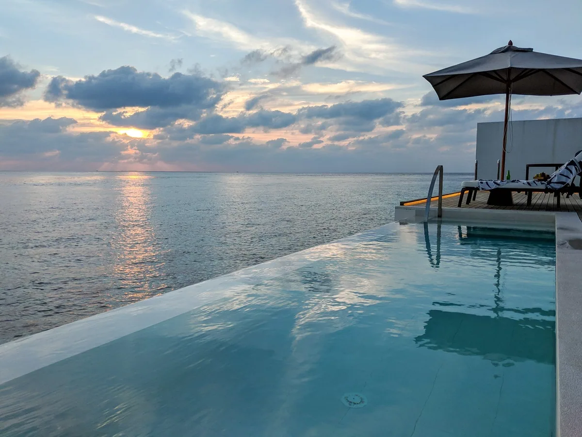 The Westin Maldives Miriandhoo Resort, villa overwater, Baa Atoll, Maldives, Overwater Pool Villa private deck at sunset, the infinity-edge plunge pool merging into the lagoon horizon, white parasol over the timber sun-lounger with the striped Westin-pattern cushion and the soft pink-and-gold sky reflecting on the still water.