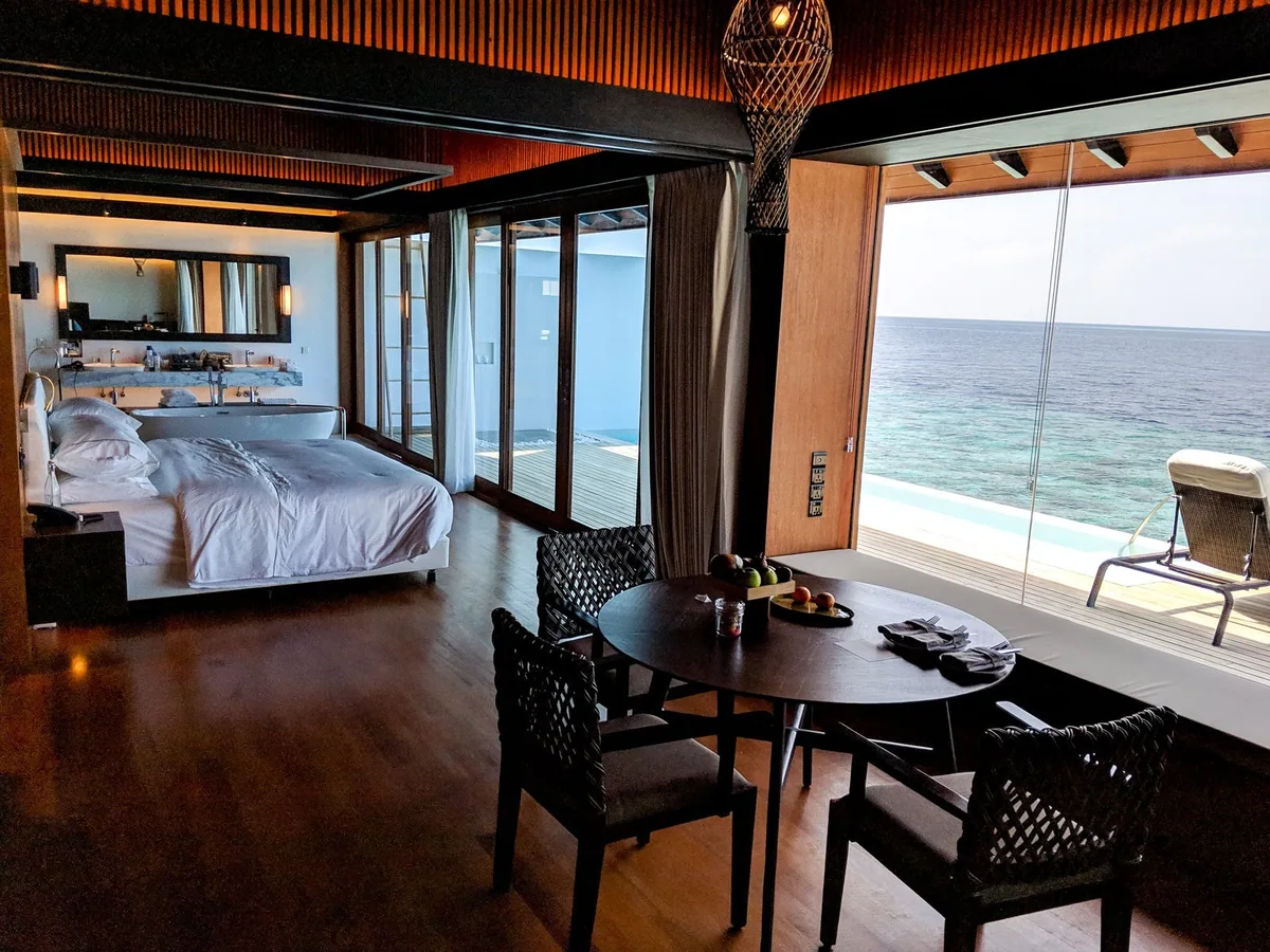 The Westin Maldives Miriandhoo Resort, villa overwater, Baa Atoll, Maldives, Overwater Pool Villa interior, the king bed with crisp white Heavenly-bed linen against the dark-slat timber feature wall, woven-rattan armchairs at the round dining table with the fruit basket, the glass-slider opening to the private deck infinity pool and the lagoon horizon.