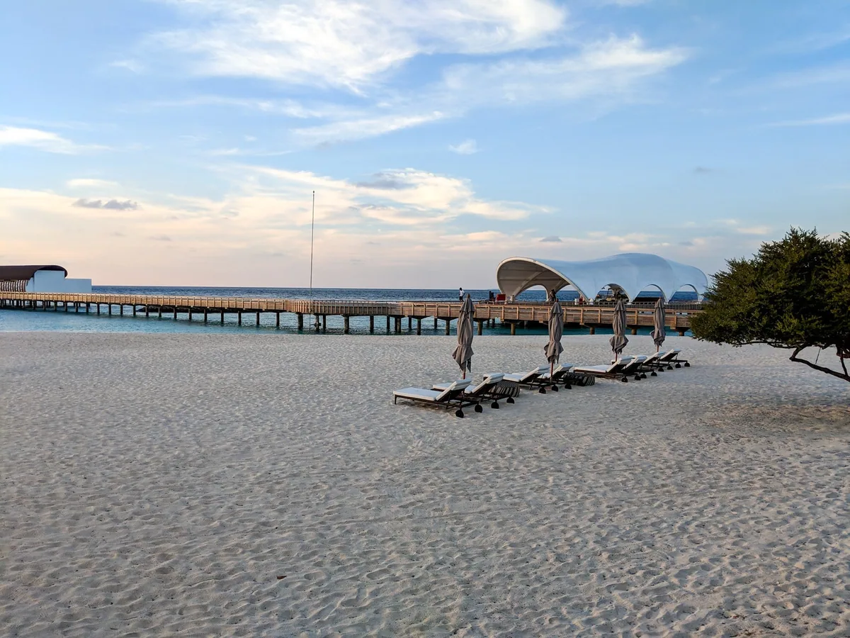 The Westin Maldives Miriandhoo Resort, setting, Baa Atoll, Maldives, The Pearl shell-shaped overwater restaurant pavilion at the end of the long timber jetty viewed from the beach, twin row of beach sun-loungers with closed cream parasols on the white sand, sea-grape tree framing the right edge under the dusk sky.
