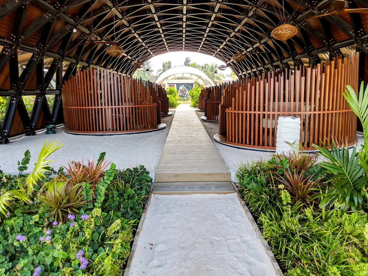 The Westin Maldives Miriandhoo Resort, spa, Baa Atoll, Maldives, Heavenly Spa by Westin entry walkway, the curved bamboo-and-timber arched tunnel with vertical-rib slatted side panels framing the path, tropical garden bursting through with crotons and ferns either side and the polished-concrete walkway leading to the spa pavilion.