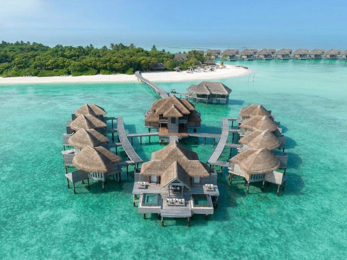 Vakkaru Maldives, spa, Baa Atoll, Maldives, Merana Spa overwater platform from above, the central spa pavilion ringed by individual thatched treatment-room pavilions connected by the curved boardwalk, the white-sand beach behind the platform and the distant overwater villa row across the lagoon.