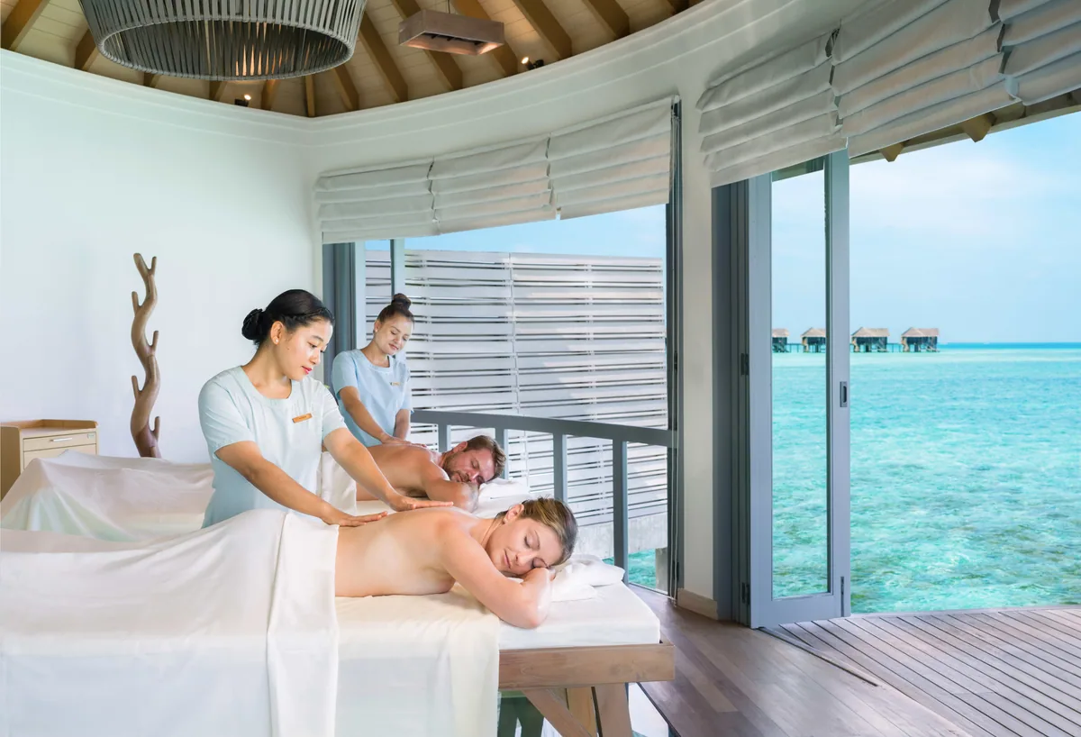 Vakkaru Maldives, spa, Baa Atoll, Maldives, Merana Spa couple's treatment room, twin guests on paired massage tables with the twin Maldivian-style therapists in pale-blue uniforms working in unison, the glass slider open to the lagoon with the overwater villa row visible across the water.