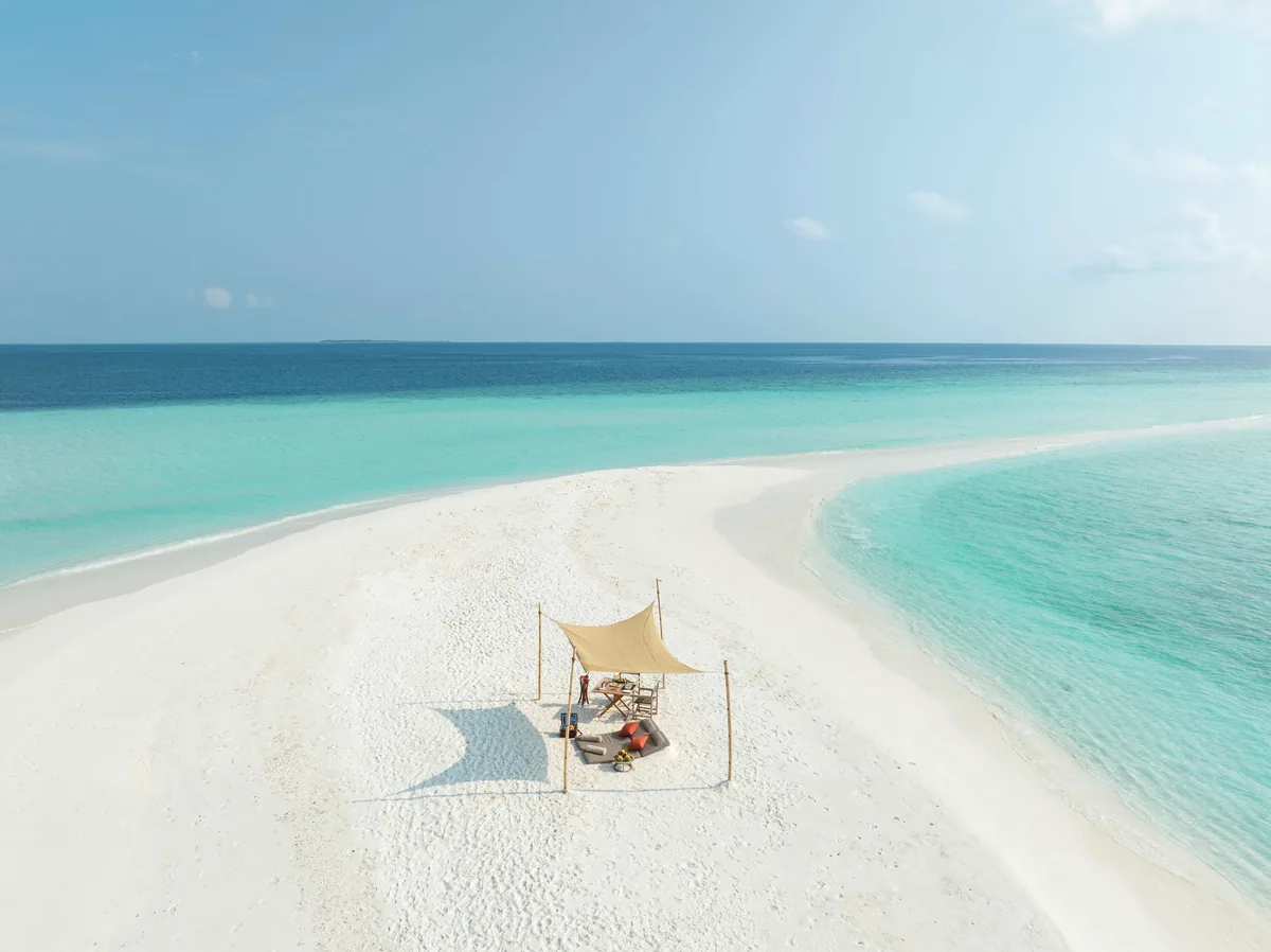 Vakkaru Maldives, activities, Baa Atoll, Maldives, Castaway sandbank private picnic setup from the drone height, the canvas sun-shade canopy with cushioned daybed and twin sun-loungers on the pristine white sandbar centred between two lagoon-blue water faces, distant atoll island silhouette on the horizon.