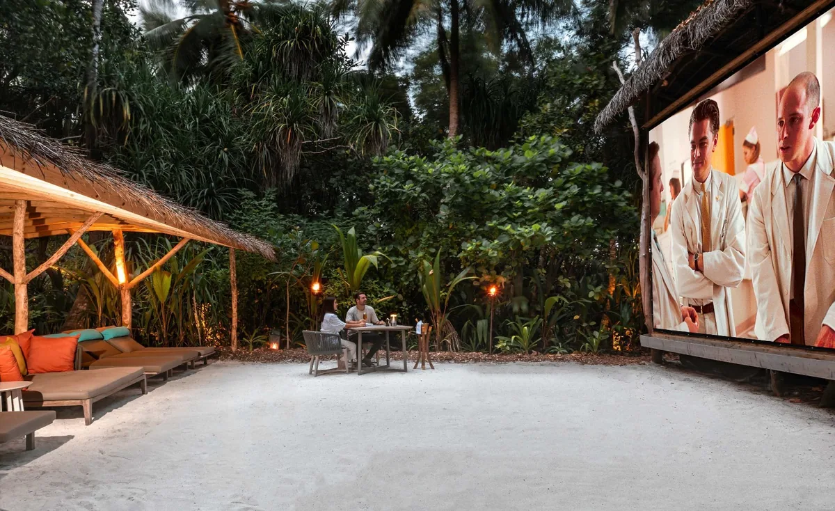 Vakkaru Maldives, activities, Baa Atoll, Maldives, Private jungle cinema after dusk, a couple at the small dining table set in the sand clearing with the large outdoor screen showing the film, orange-and-teal cushioned sun-loungers on the thatched cabana to the left, dense tropical jungle framing the open-air theatre.