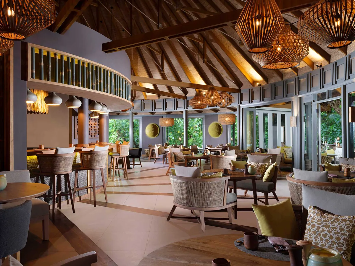 Vakkaru Maldives, dining, Baa Atoll, Maldives, Onu Southeast-Asian restaurant interior, the multi-tier dining hall with bamboo-woven pendant lanterns and the central bamboo lampshade chandelier, modern slate-and-yellow chair palette, walk-around bar with mirror back, lush jungle visible through the floor-to-ceiling glass.