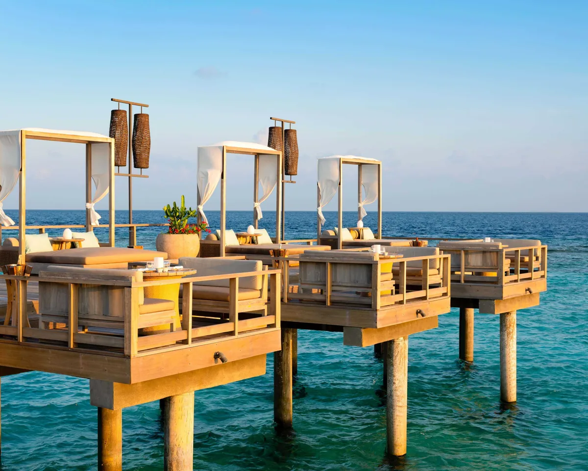 Vakkaru Maldives, dining, Baa Atoll, Maldives, Lagoon Bar at golden hour, three stilted overwater platform daybed pavilions in formation, each with sheer cream canopy curtains and woven-rattan pendant lanterns over the cushioned timber daybeds, the soft warm light reflecting on the turquoise lagoon.
