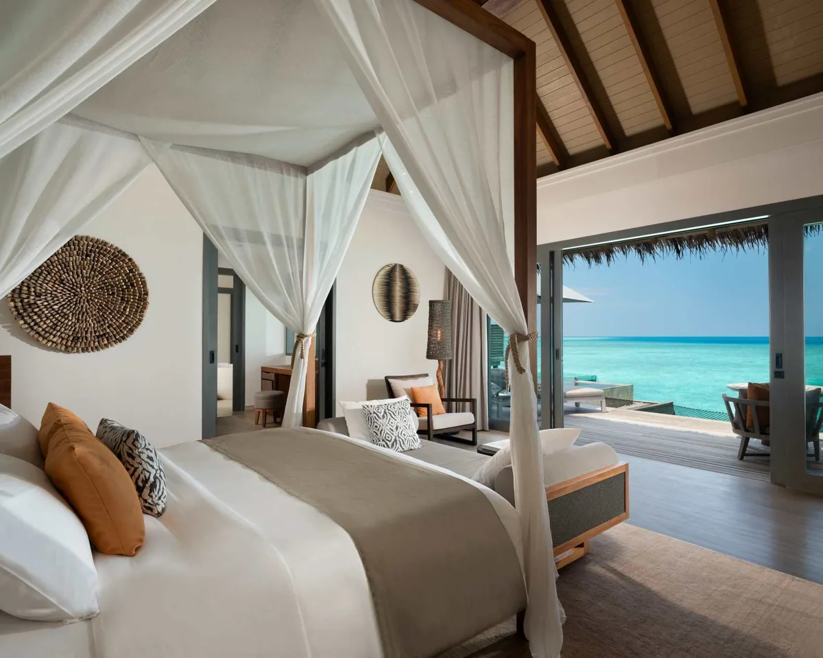 Vakkaru Maldives, villa overwater, Baa Atoll, Maldives, One-Bedroom Overwater Pool Residence bedroom, the four-poster timber bed with sheer cream canopy, terracotta-orange and zebra-pattern cushions, large woven-disk wall art beside the woven-globe pendant lantern, glass doors opening to the private deck pool and lagoon.