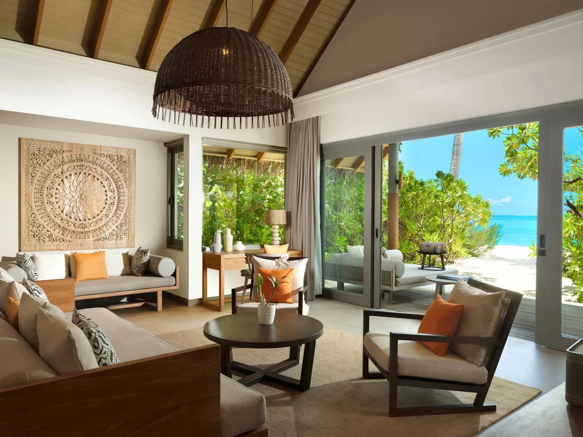 Vakkaru Maldives, villa beach, Baa Atoll, Maldives, One-Bedroom Beach Pool Residence living room, the large carved-Maldivian-wood medallion above the cream sofa with orange-and-zebra accent cushions, woven-globe pendant overhead, glass sliders opening directly to the sea-grape-tree-framed deck and beach.