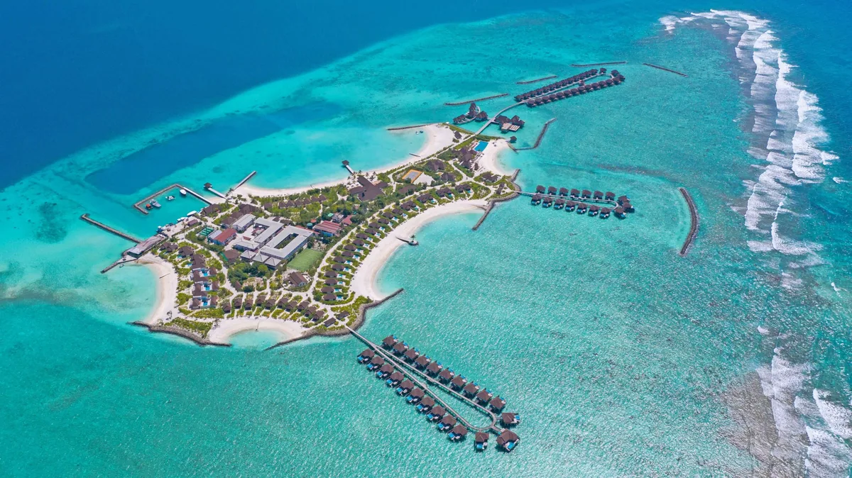 VARU by Atmosphere, aerial, North Malé Atoll, Maldives, Full property aerial in clear midday light, the arrowhead-shape island with the central main service block and beach villa cluster, twin U-shape over-water villa jetties extending toward the reef at the right (the sunrise and sunset Water Villa configurations), and the perimeter reef breakline against the deeper ocean.