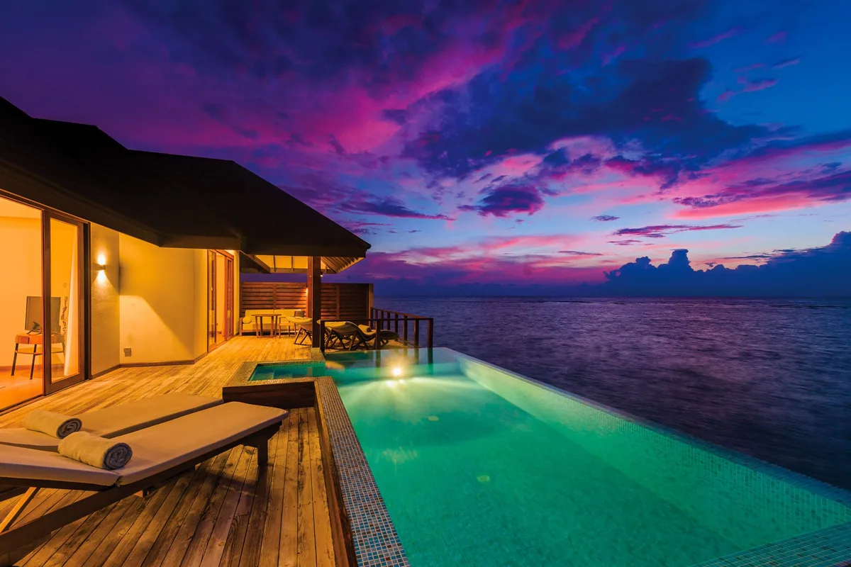 VARU by Atmosphere, villa overwater, North Malé Atoll, Maldives, Water Suite exterior at twilight, the standalone over-water villa with private infinity pool extending toward the lagoon edge, sun loungers and lounge chairs on the timber deck, warm interior light spilling through the sliding glass walls, and a magenta-purple-blue Maldivian sunset sky overhead.