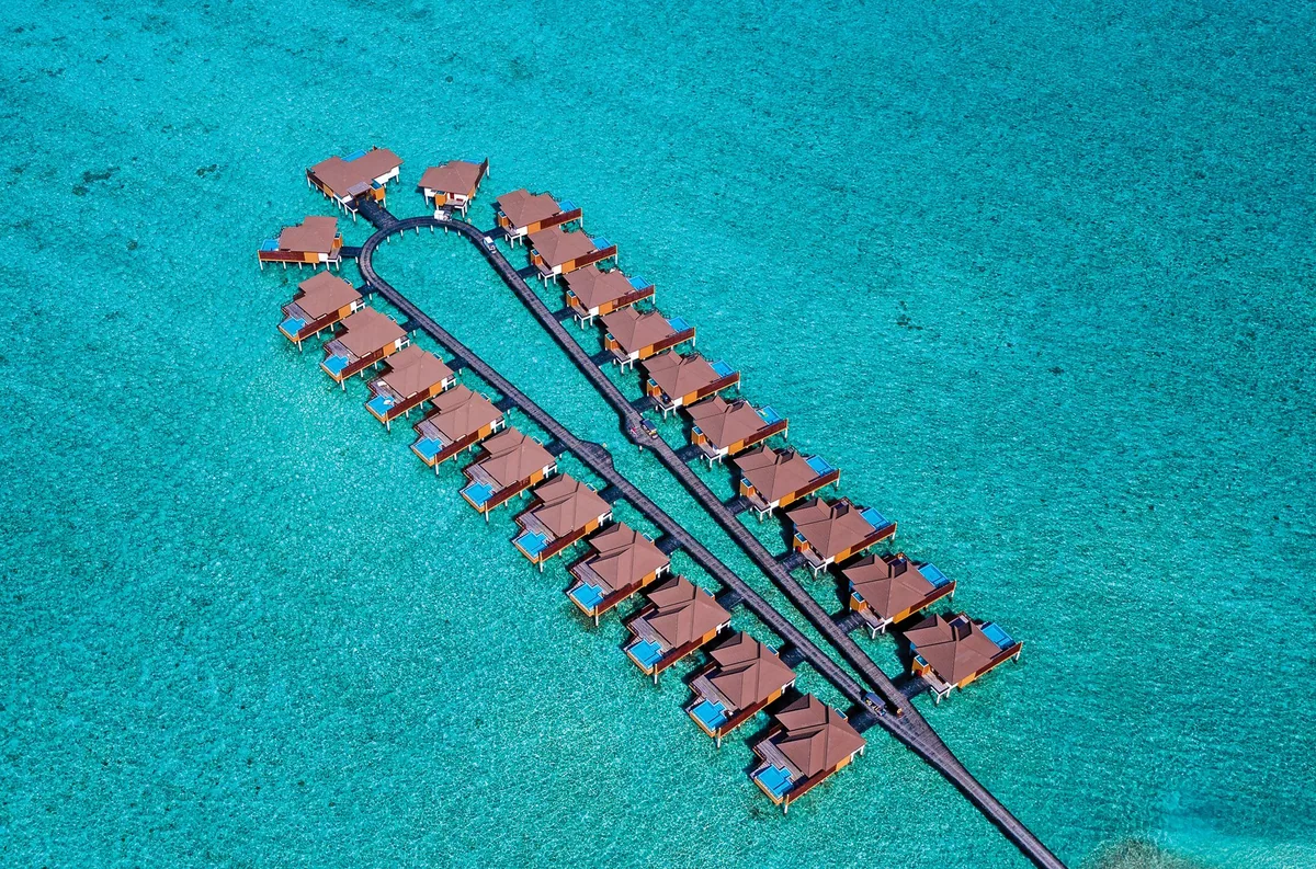 VARU by Atmosphere, villa overwater, North Malé Atoll, Maldives, Aerial of the over-water villa jetty cluster, the U-shape twin-boardwalk configuration with the brick-red pitched-roof villas tracking the parallel jetties across the crystal turquoise reef shallows (the property's signature Water Villa geometry).