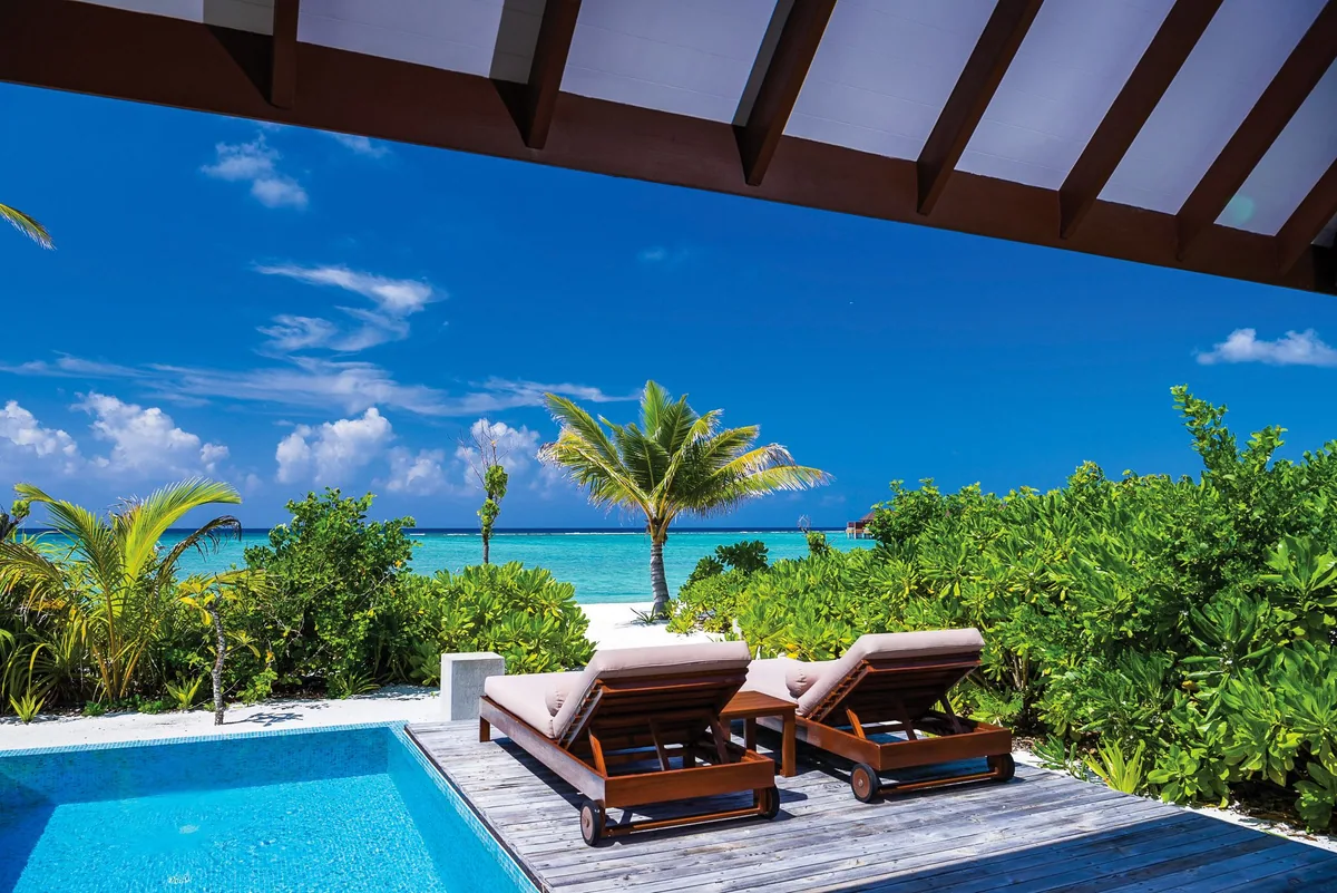 VARU by Atmosphere, villa beach, North Malé Atoll, Maldives, Beach Villa with Pool deck, the small private rectangular plunge pool at the deck edge, two cushioned wooden sun loungers facing the lagoon, palm canopy framing the view to the powder-white perimeter beach, and dark-stained timber decking.