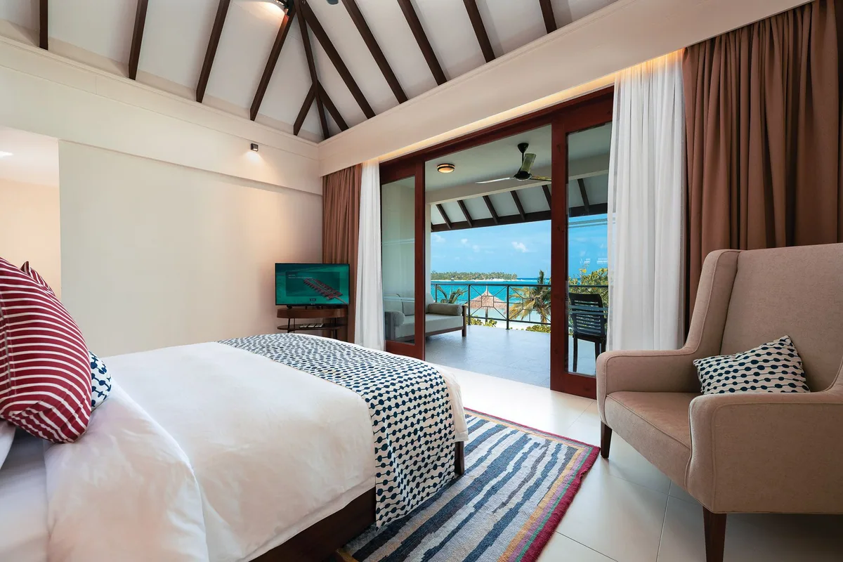 VARU by Atmosphere, villa beach, North Malé Atoll, Maldives, Majlis suite master bedroom interior, the high pitched ceiling with dark-stained timber rafter structure, a tall cream-upholstered armchair, red-and-cream striped throw cushions, a blue-and-cream geometric throw across the bed, the woven striped runner rug, and the open sliding doors framing the veranda with ocean view.