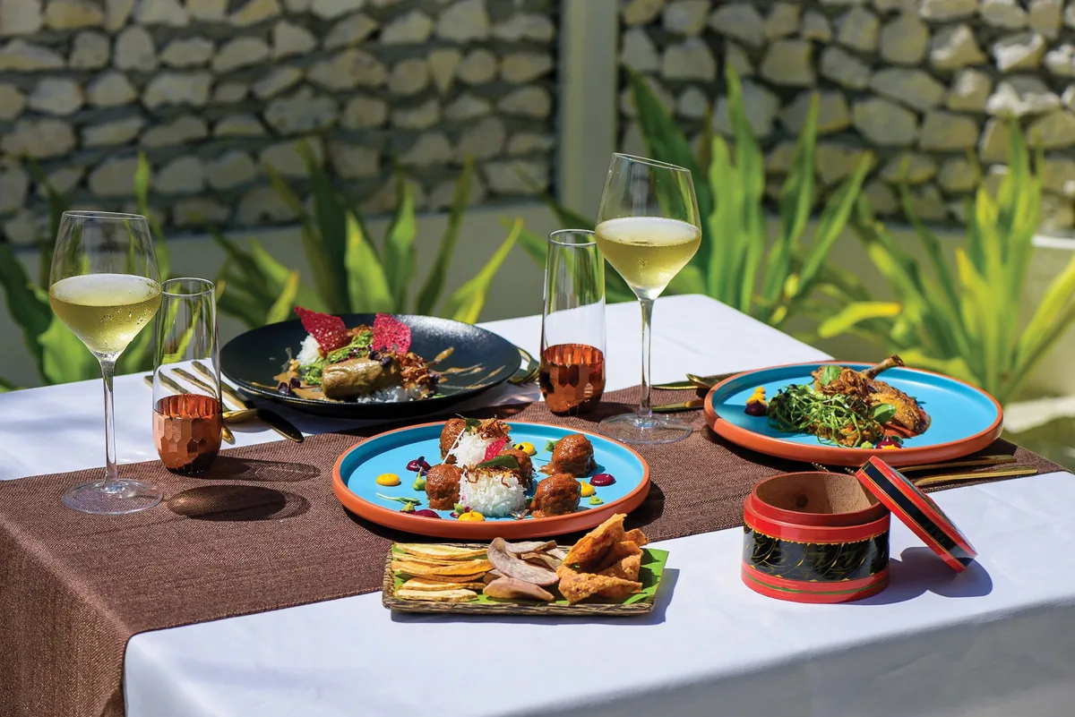 VARU by Atmosphere, dining, North Malé Atoll, Maldives, Kaage Maldivian-cuisine plating, the orange-and-turquoise stoneware plate with a contemporary preparation of Maldivian meatballs and rice garnished with herbs and edible flowers, a traditional lacquered Maldivian condiment box, gold-tone cutlery, copper goblets, and the coral-stone wall backdrop with tropical foliage.